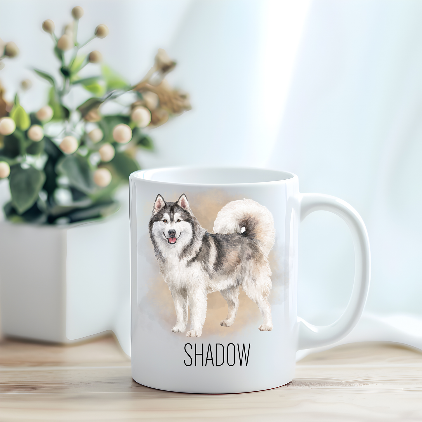 This coffee mug features a Husky on a white ceramic mug which has been placed on a light wooden surface.  A green and white plant sits on the left side of the image in a square plant pot.