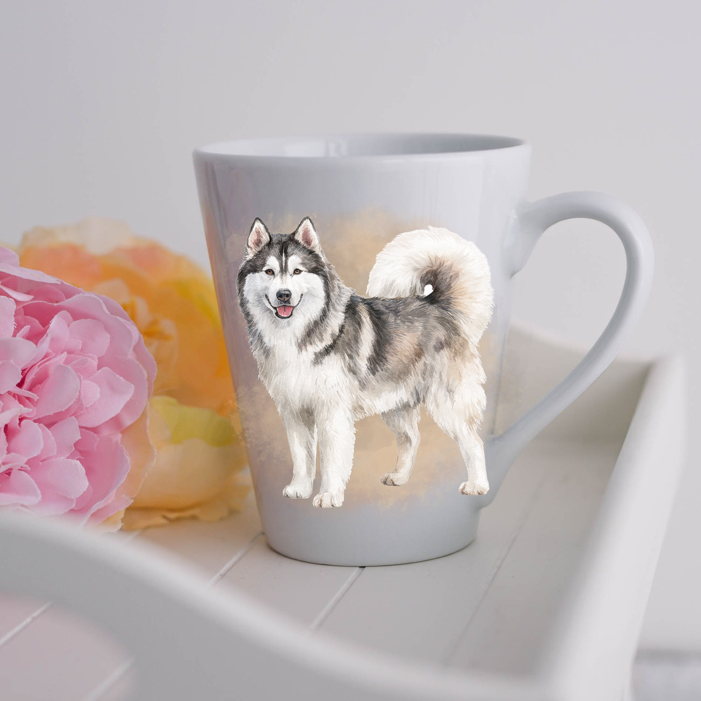 A Husky is shown standing on a white ceramic latte mug in a smoky background and looking forward, mouth open as if smiling.  The mug is placed on a white tray with pink and yellow flowers. 
