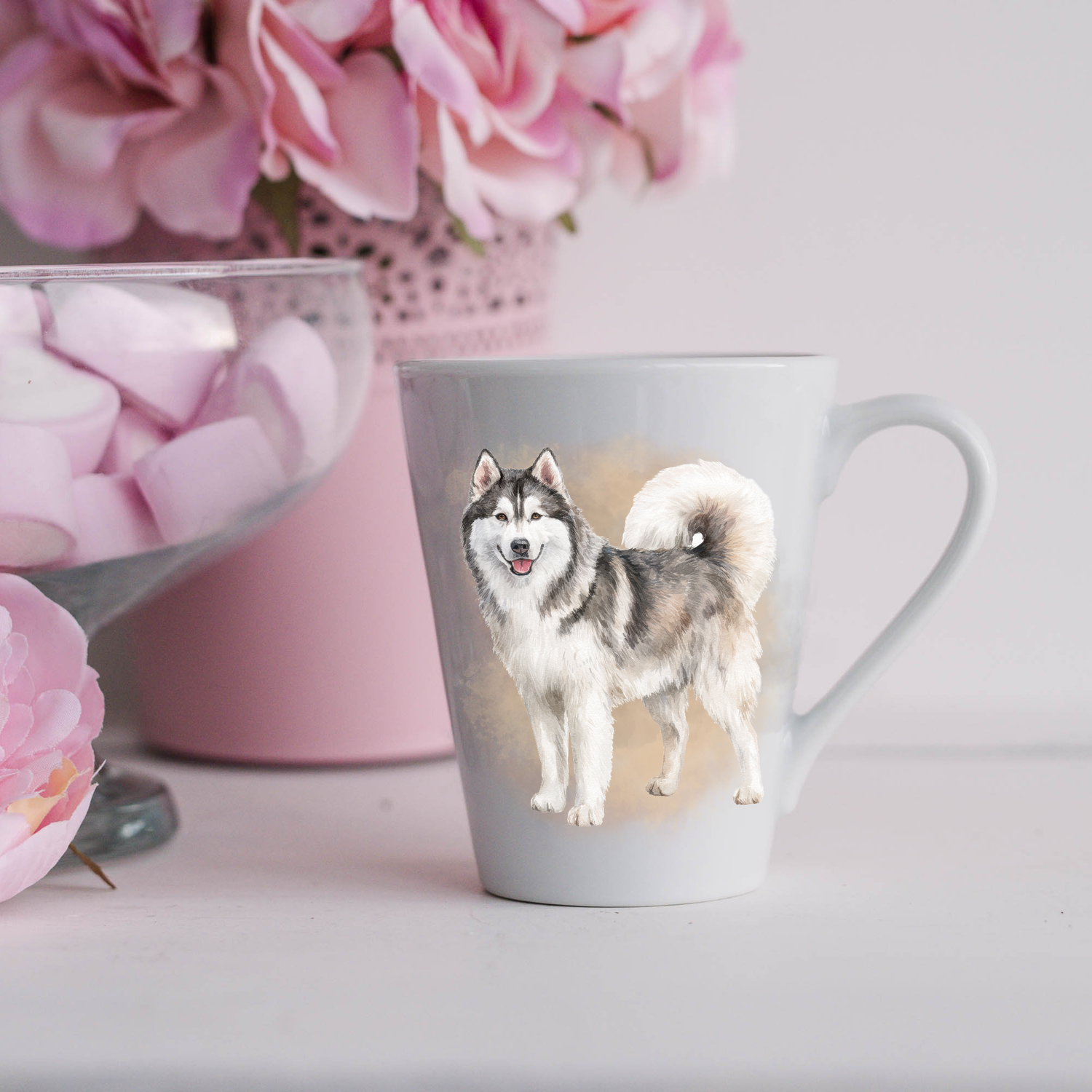 A Husky is shown standing on a white ceramic latte mug in a smoky background and looking forward.  He has a smile on his face and a bushy tail curled over his back.  The mug is placed on a white surface with pink flowers in a pink plant pot and a dish of pink marshmallows.