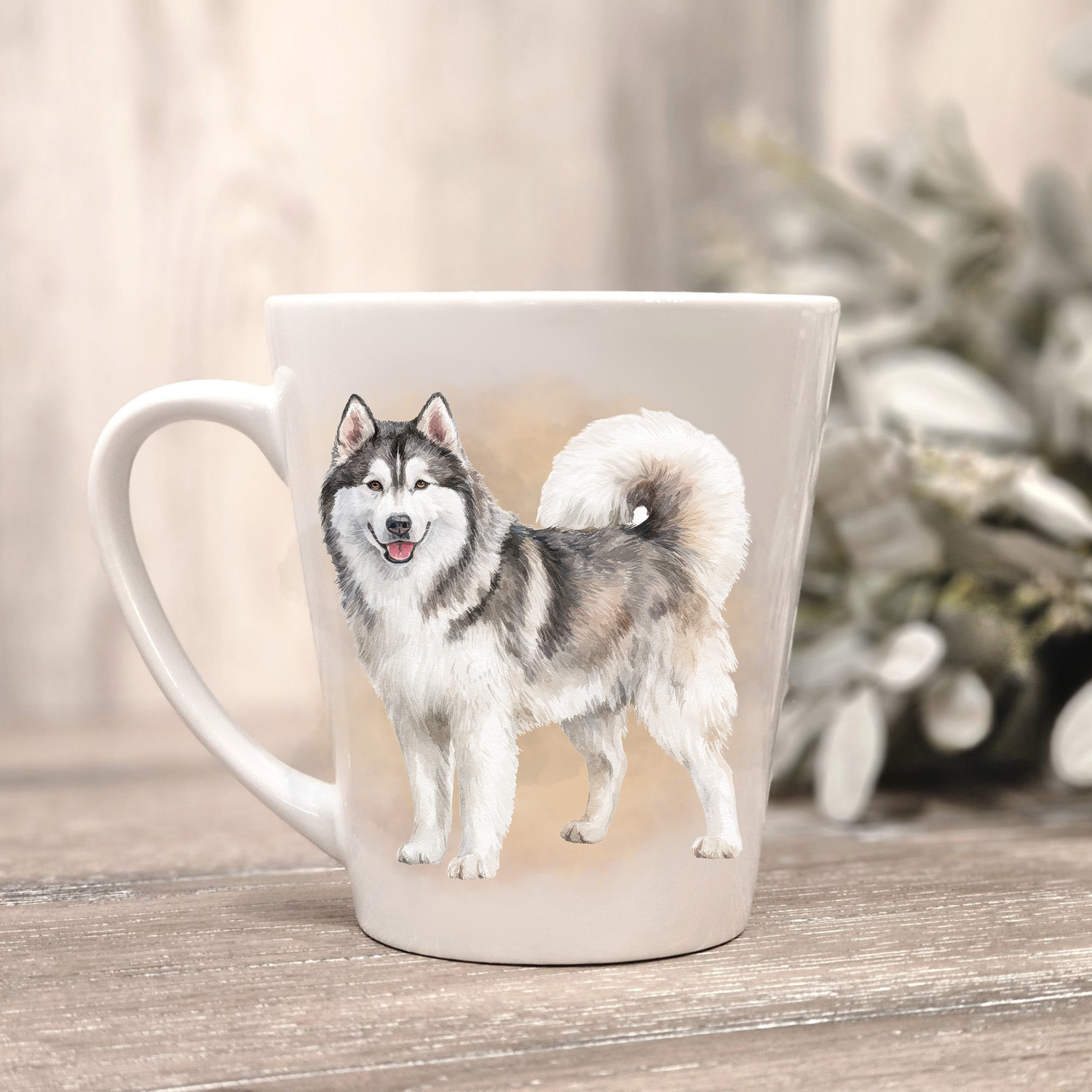 A Husky is standing on a white ceramic latte mug in a smoky background and looking forward.  The mug is placed on a wooden surface with sage green foliage to the right-hand side.