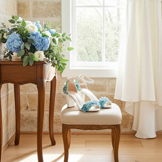 Pair of high-heeled bridal shoes with floral decorations on a chair next to a table with flowers.