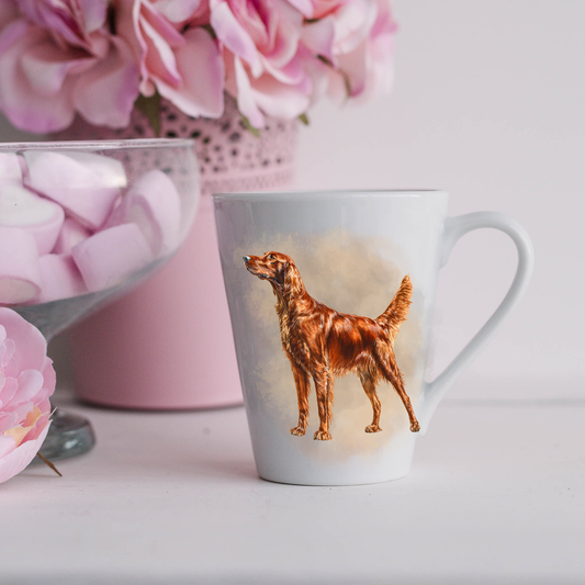 A beautiful Irish Setter is seen standing on a white ceramic latte mug in a smoky background and looking to his right.  The mug is placed on a white surface with pink flowers in a pink plant pot and a dish of pink marshmallows.