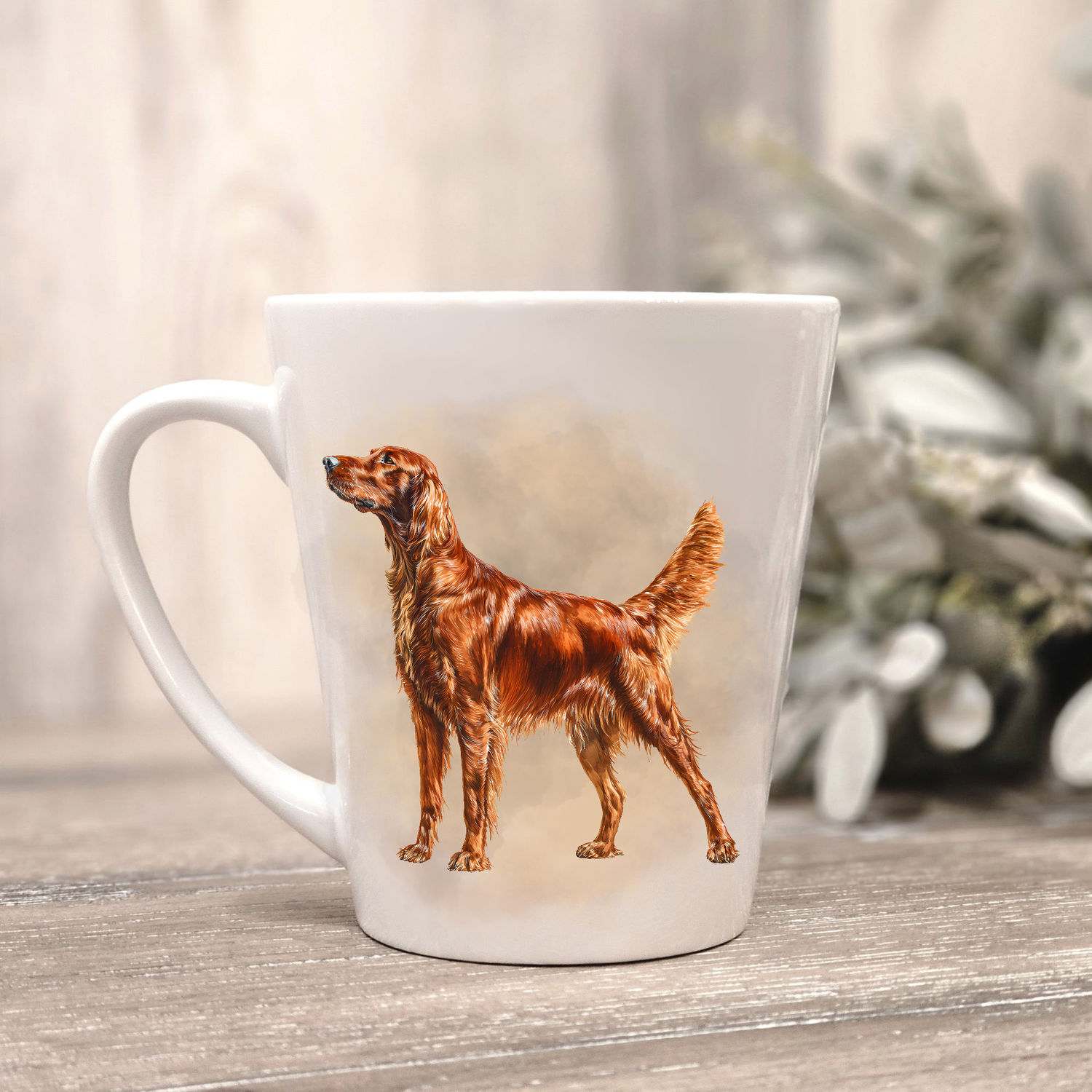 An Irish Setter is seen standing on a white ceramic latte mug in a smoky background and looking forward.  The mug is placed on a wooden surface with sage green foliage to the right-hand side.