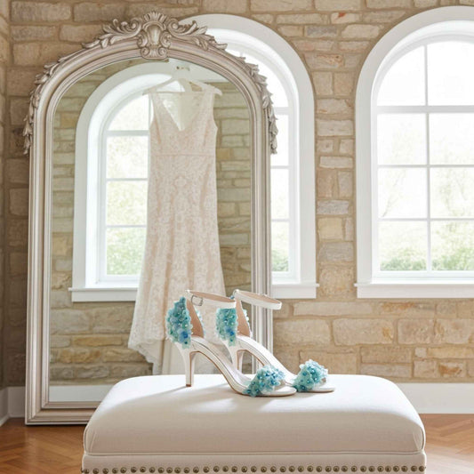 Ivory high-heeled shoe with blue floral decorations on a white ottoman in front of a large mirror reflecting a wedding dress.