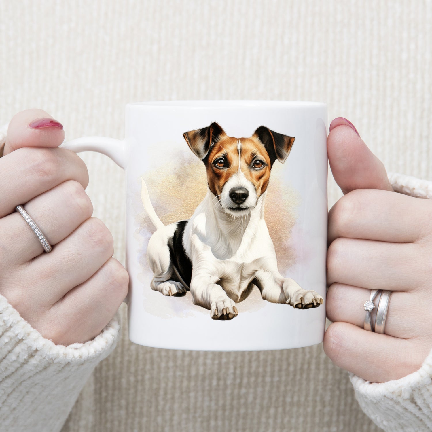 A woman is holding white ceramic mug showing a Jack Russell Terrier laying in front of a brown and gold misty background and facing forward.