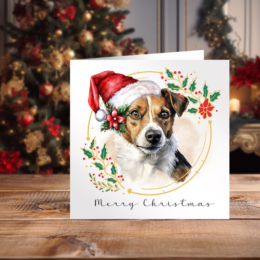 Jack Russell Terrier wearing a Santa hat on a white Christmas themed card positioned on a wooden surface in front of a Christmas tree.