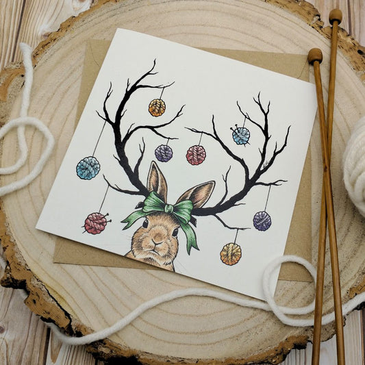 A greetings card featuring an illustration of a rabbit with antlers, decorated with hanging wool baubles, on a wooden surface with a wooden round panel in the background. The card also shows a portion of wool and a knitting loom in the foreground.