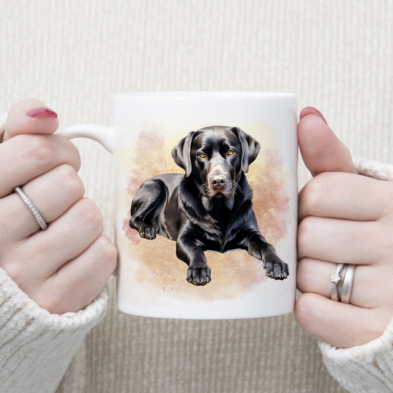 A black Labrador dog is laid among a pink and gold misty background on a  white ceramic mug. A woman is holding the mug