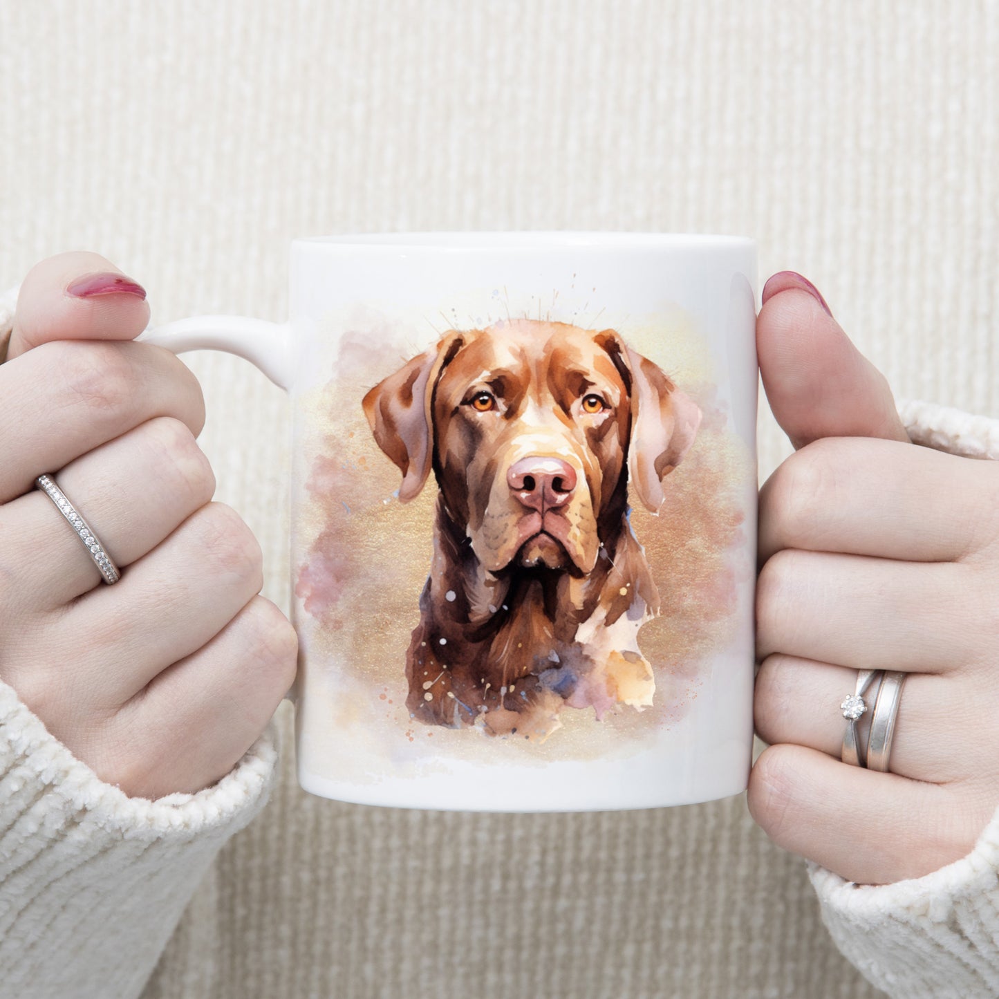 A watercolour image of a Chocolate Labrador dog adorns this white ceramic mug.  A pink and gold misty background decorates the mug which is being held by a woman with both hands.