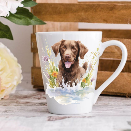 A white ceramic latte coffee mug with a watercolor design of a chocolate Labrador dog, surrounded by floral elements, placed on a wooden surface with a floral background.