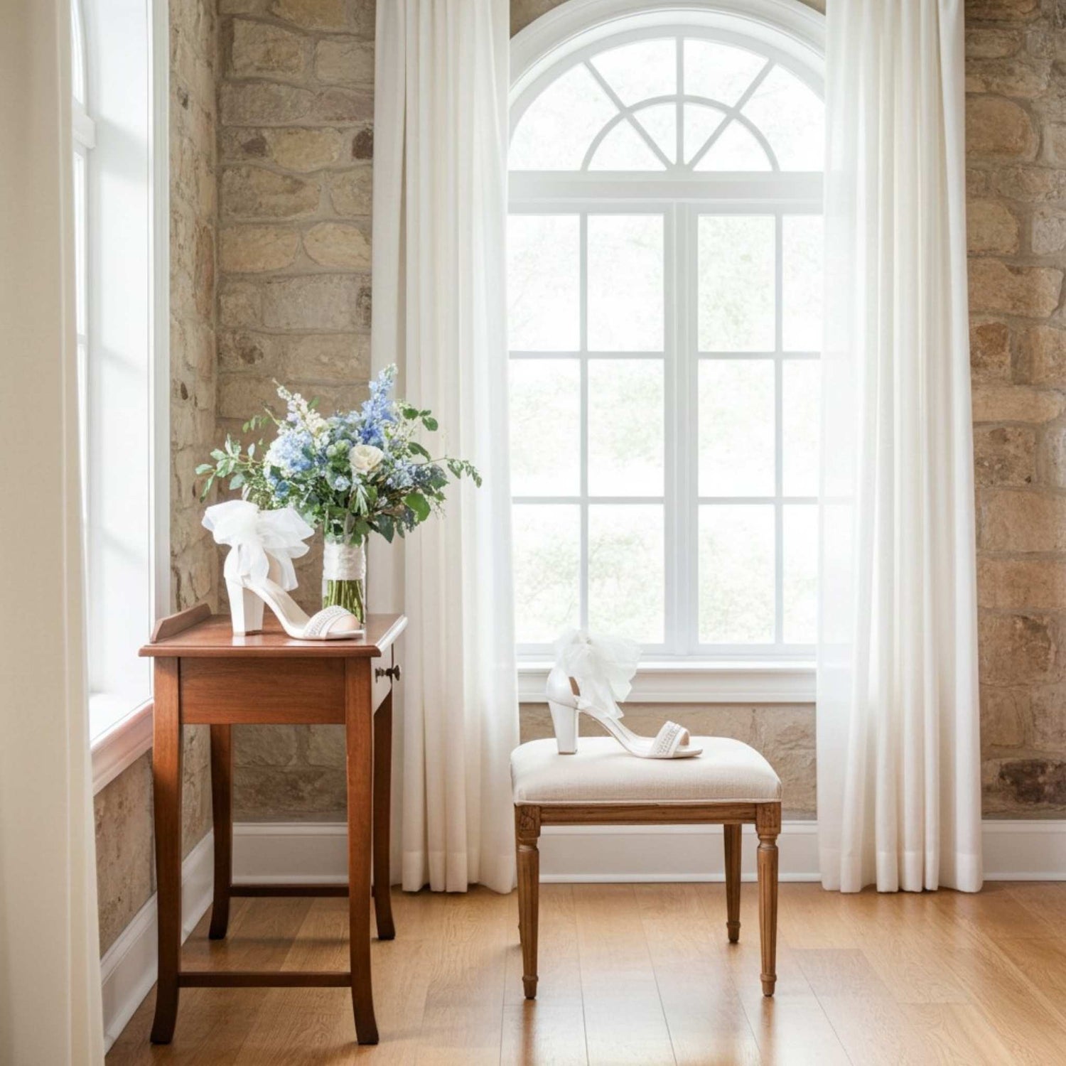 Wooden table with a vase of flowers and ivory block heel bridal shoes, next to a cushioned stool in a bright room with stone walls and large windows.