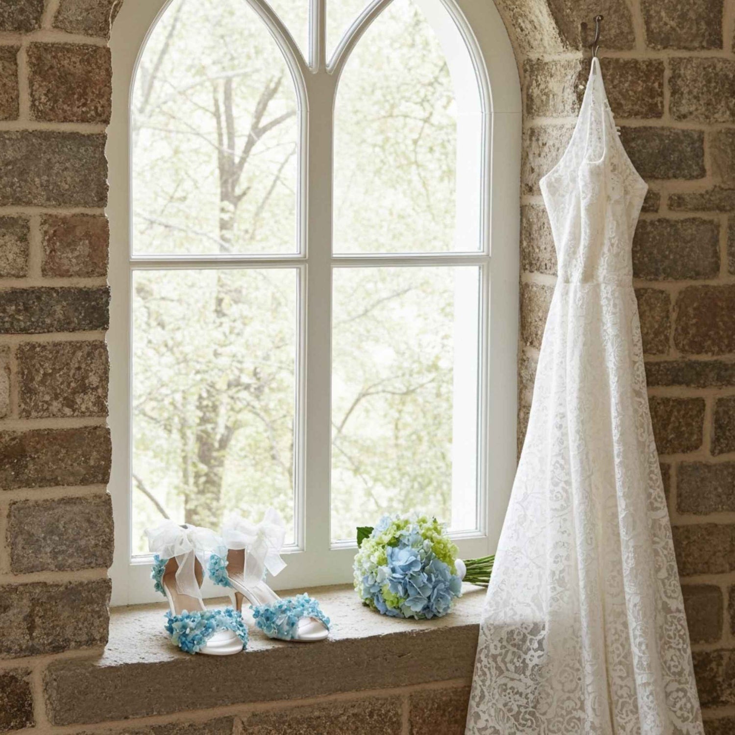 White wedding dress hanging on a brick wall with bridal shoes and flowers on a windowsill.