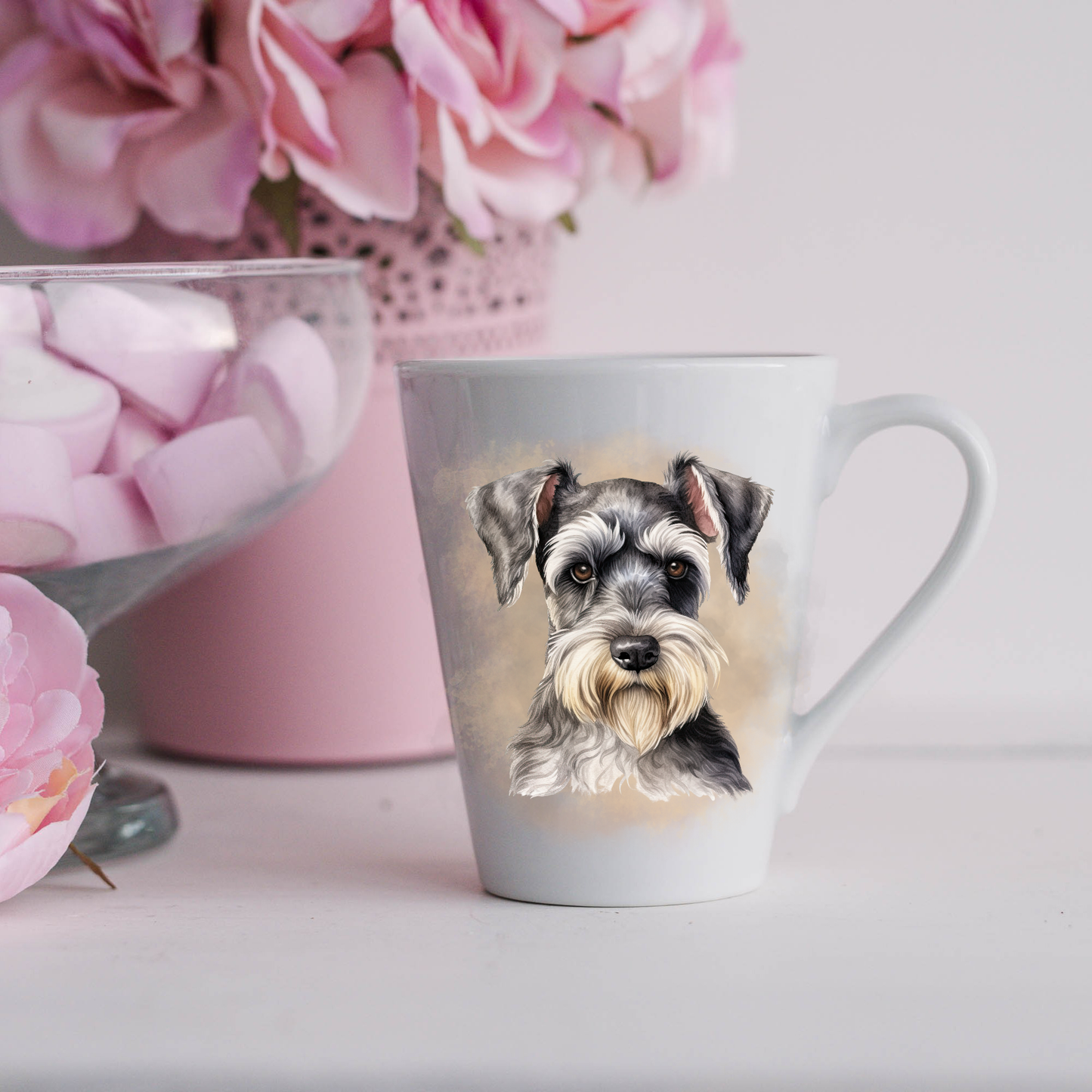 A Miniature Schnauzer is shown on a white ceramic latte mug in a smoky background and looking forward.  The mug is placed on a white surface with pink flowers in a pink plant pot and a dish of pink marshmallows.