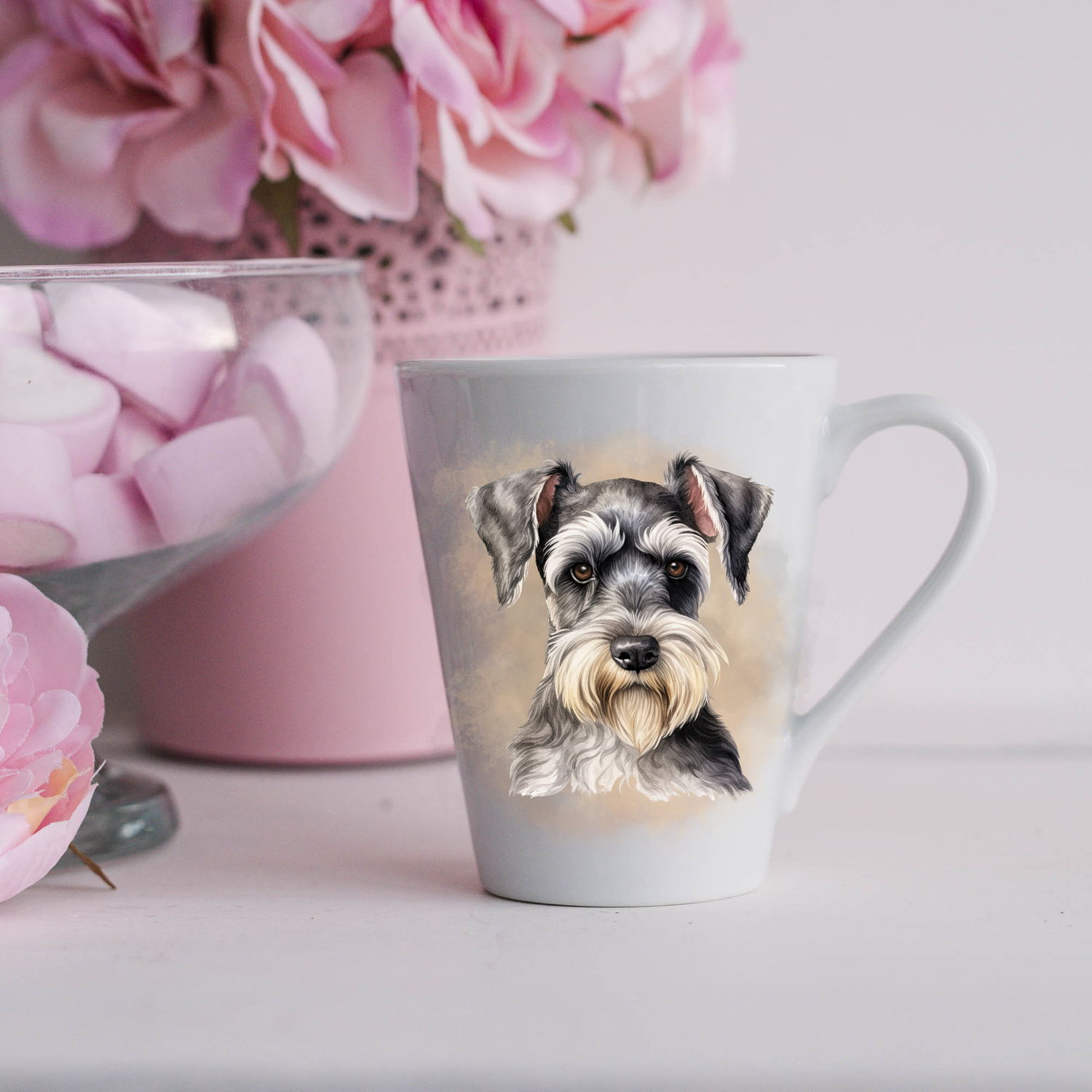 A Miniature Schnauzer is shown on a white ceramic latte mug in a smoky background and looking forward.  The mug is placed on a white surface with pink flowers in a pink plant pot and a dish of pink marshmallows.