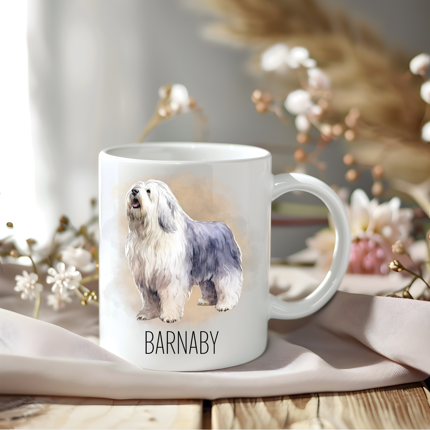 An Old English Sheepdog coffee mug personalised with a pet name.  The mug is sat on a wooden surface with a creamy coloured cloth and various flowers adorn the background.