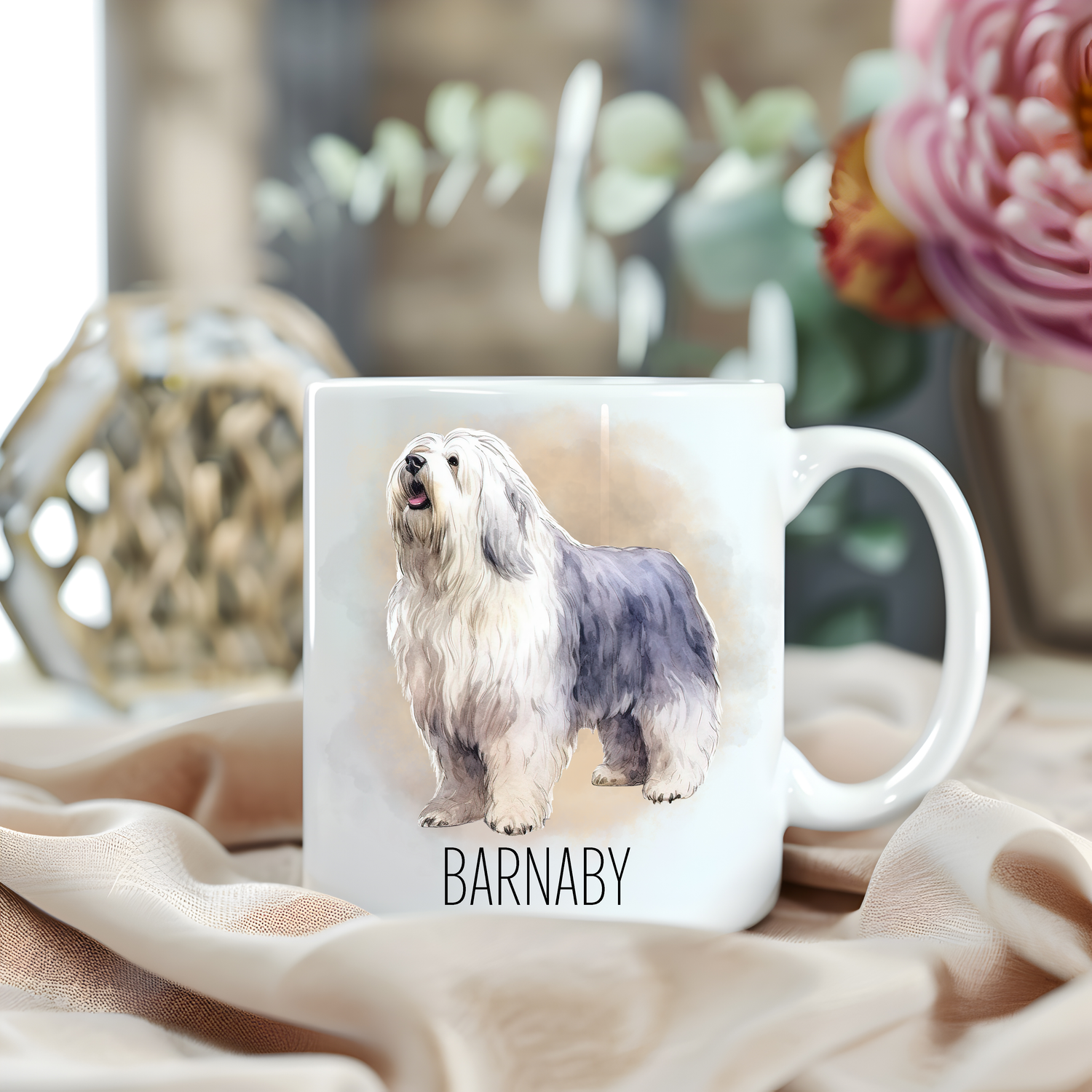An Old English Sheepdog is stood looking to the left on a white personalised ceramic mug which has been placed on a beige cloth.  A large pink flower with light green foliage accessorizes the image.