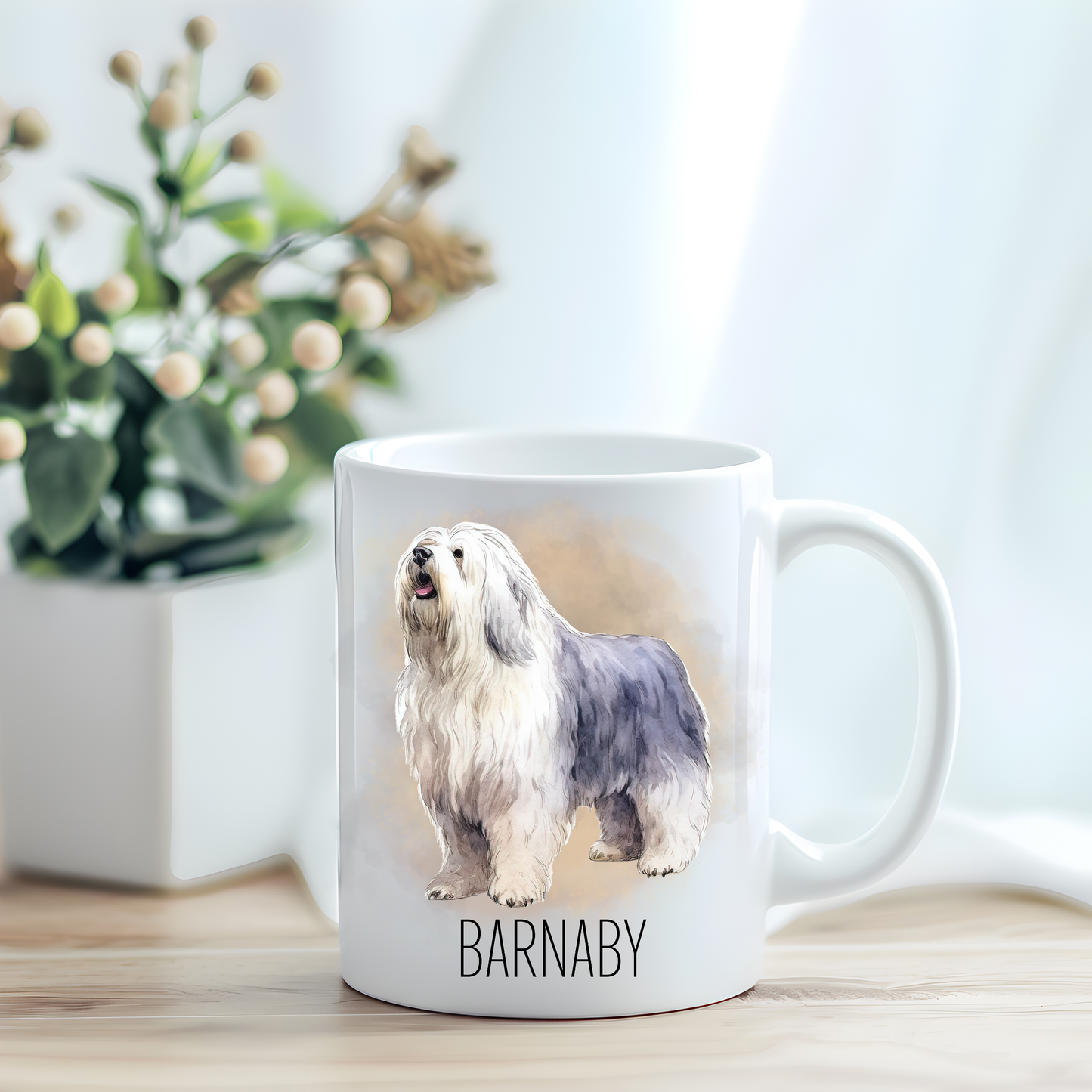 This coffee mug features an Old English Sheepdog on a white ceramic mug which has been placed on a light wooden surface.  A green and white plant sits on the left side of the image in a square plant pot.
