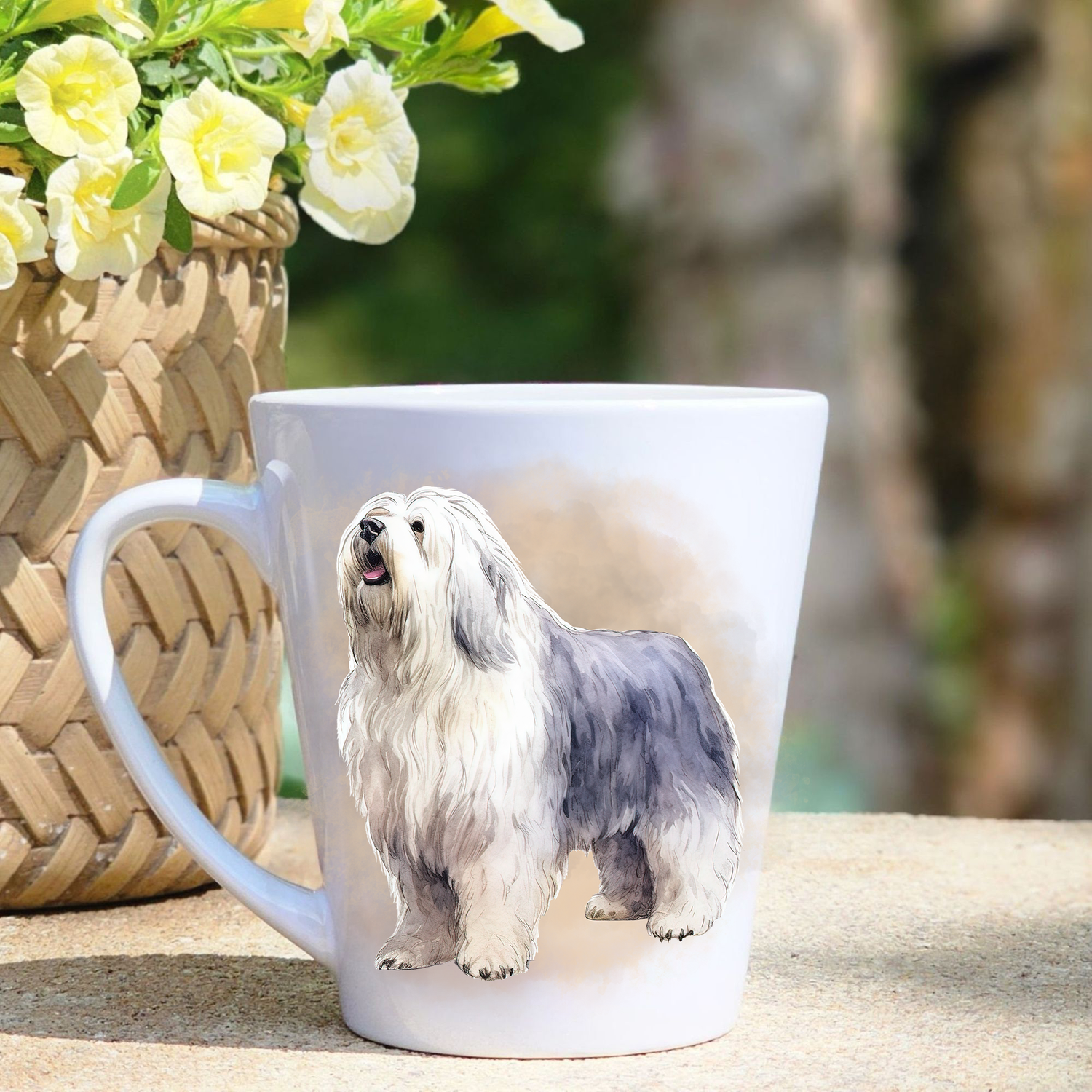 A white ceramic latte mug featuring a standing Old English Sheepdog in a beige mist background.  A basket containing yellow flowers sits to the left-hand side.