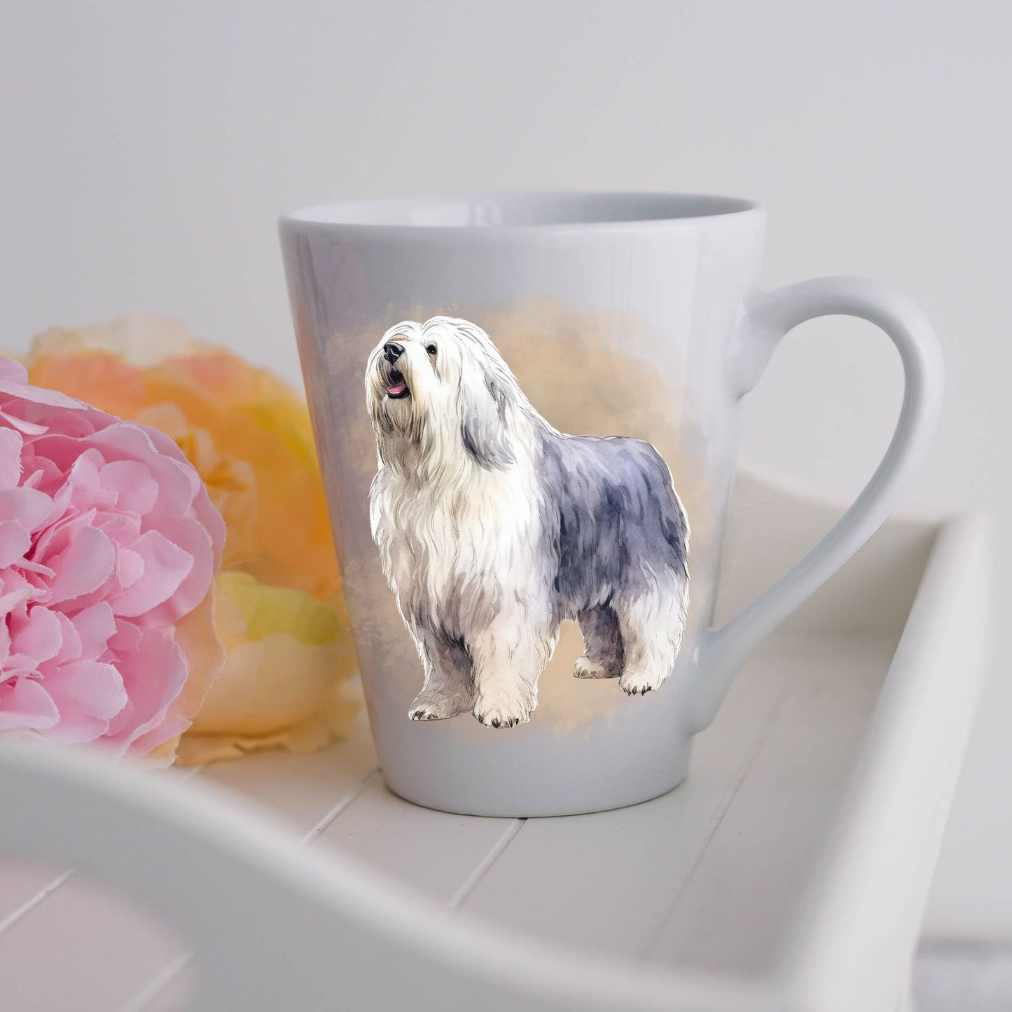 An Old English Sheepdog is shown standing on a white ceramic latte mug in a smoky background and looking up and to his left.  The mug is placed on a white tray with pink and yellow flowers. 