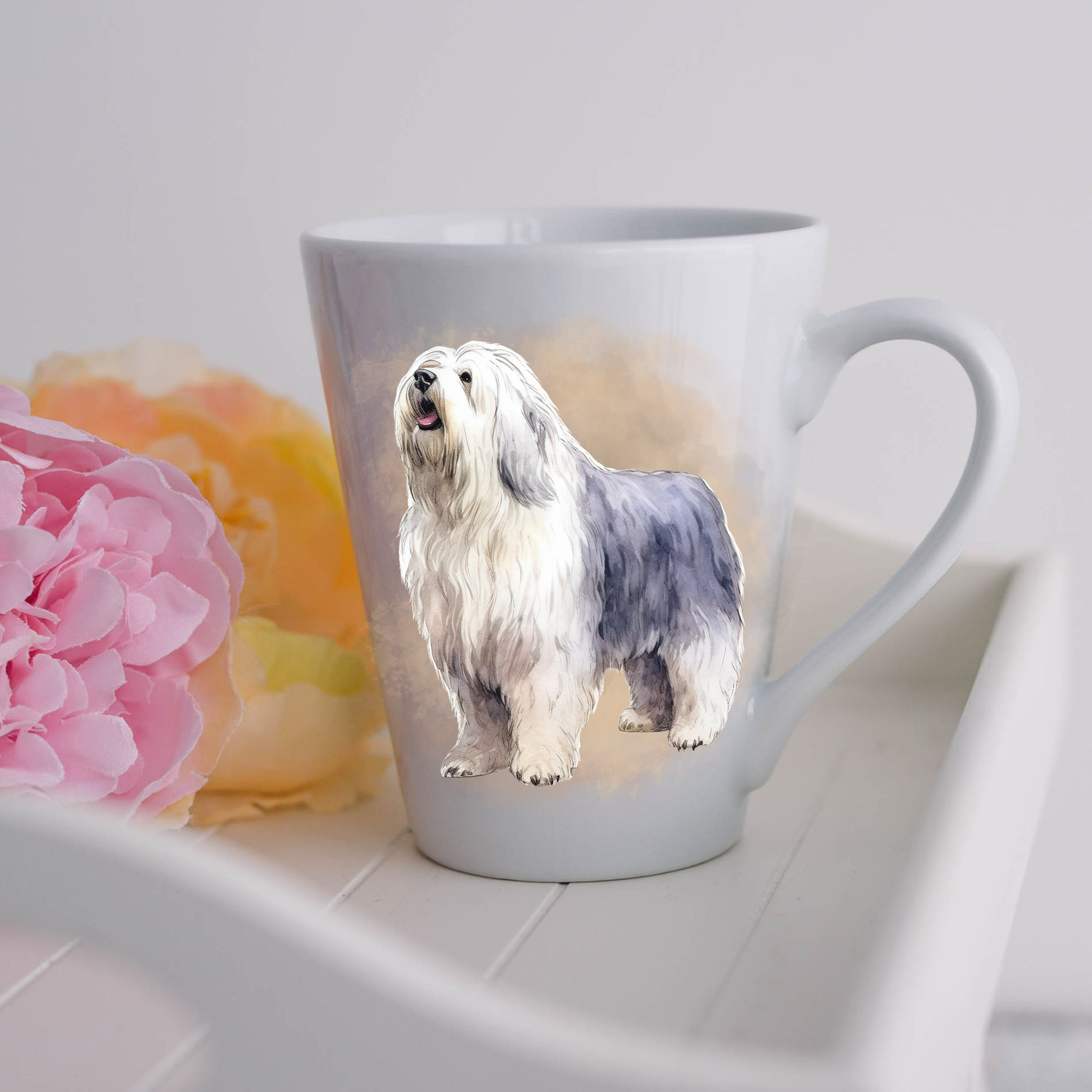 An Old English Sheepdog is shown standing on a white ceramic latte mug in a smoky background and looking up and to his left.  The mug is placed on a white tray with pink and yellow flowers. 