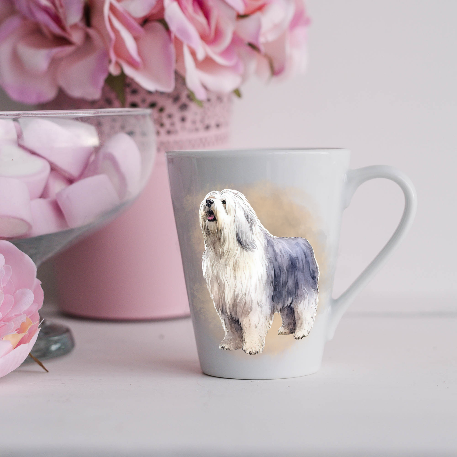 An Old English Sheepdog is seen standing on a white ceramic latte mug in a smoky background and looking forward.  The mug is placed on a white surface with pink flowers in a pink plant pot and a dish of pink marshmallows.