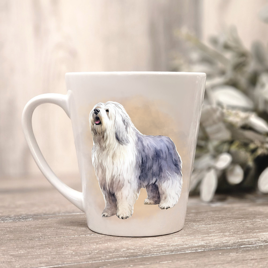 An Old English Sheepdog is shown standing on a white ceramic latte mug in a smoky background and looking forward.  The mug is placed on a wooden surface with sage green foliage to the right-hand side.
