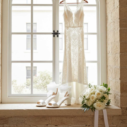 Ivory wedding dress, low block heel bridal shoes, and bouquet on a windowsill with a brick wall background