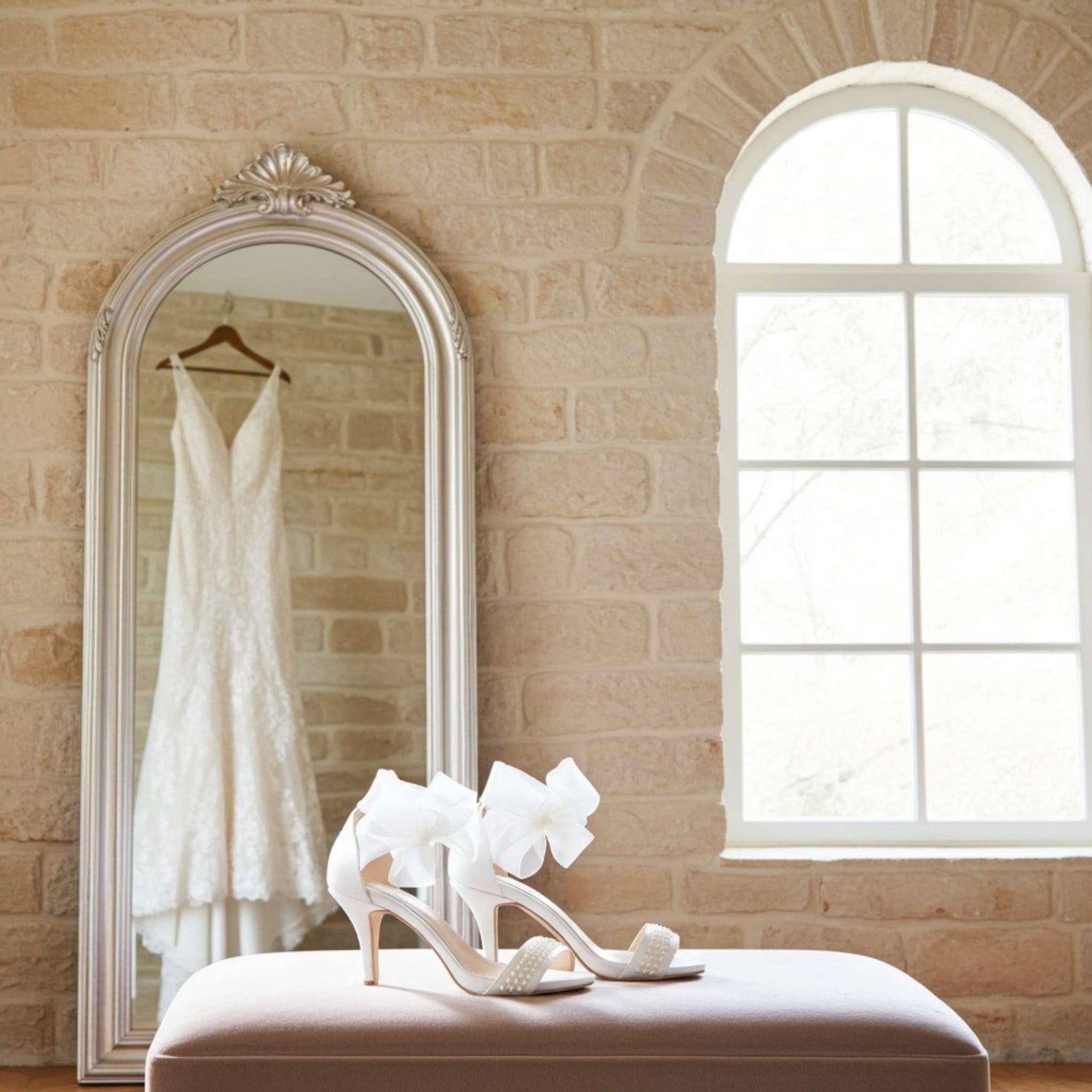 White wedding dress hanging in a mirror with ivory high-heeled pearl wedding shoes on a bench in a room with a brick wall and window.