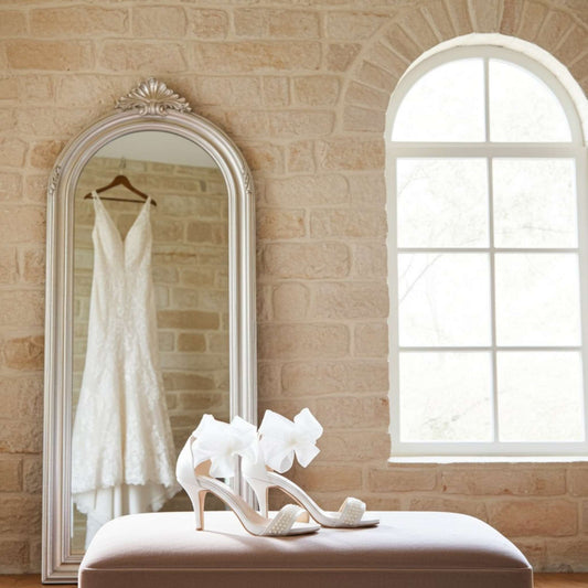 White wedding dress hanging in a mirror with ivory high-heeled pearl wedding shoes on a bench in a room with a brick wall and window.