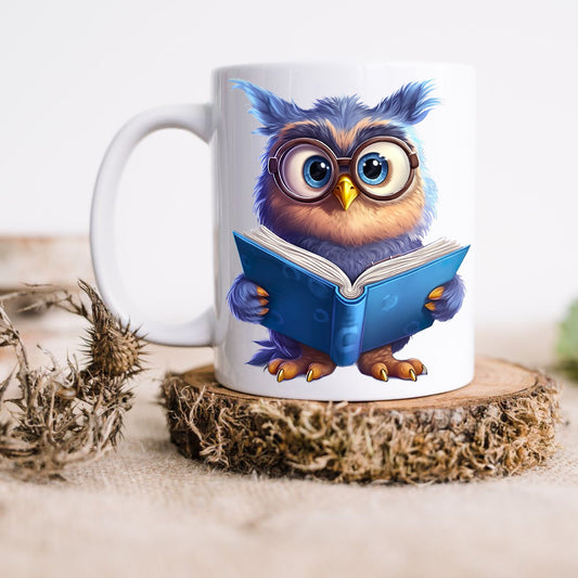A blue Owl wearing glasses is reading a blue book on a white ceramic mug. The mug is placed on a thick wooden disc and a dried thistle is set to the left hand side.