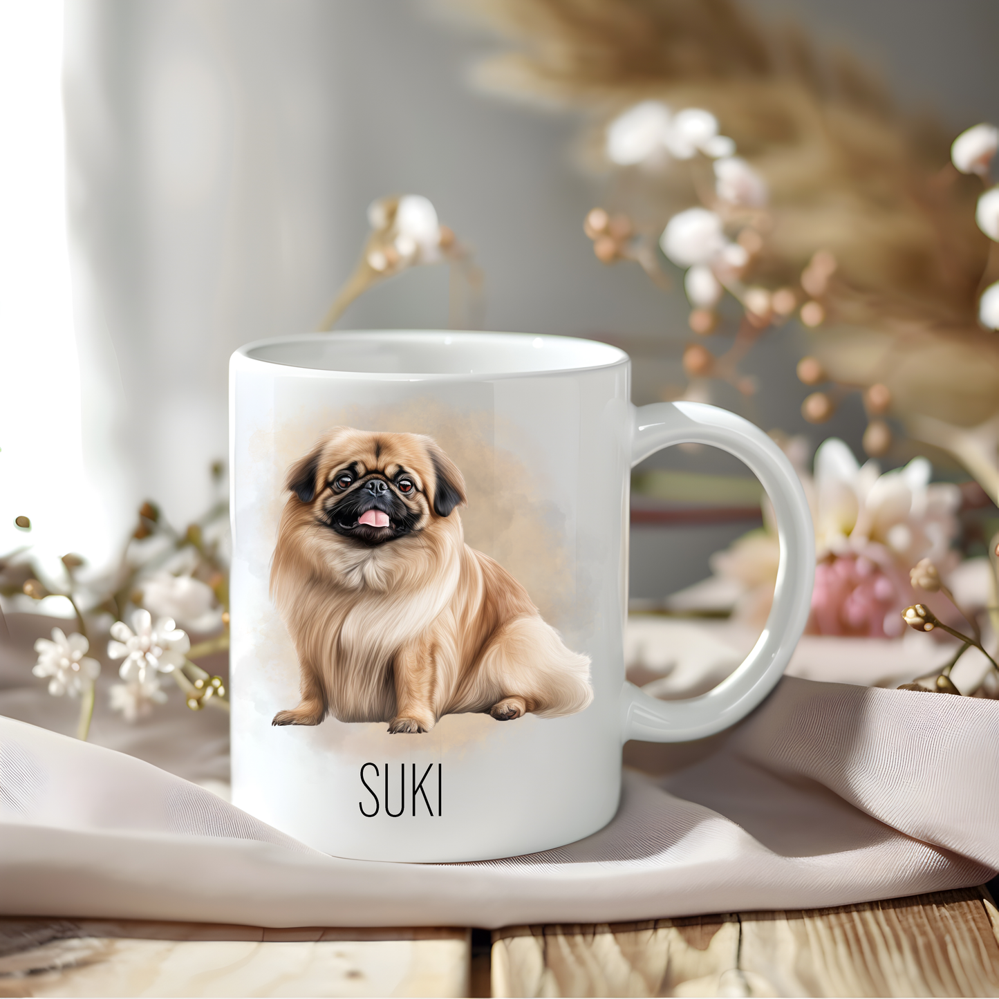 A Pekingese dog coffee mug personalised with a pet name.  The mug is sat on a wooden surface with a creamy coloured cloth and various flowers of pink and white adorn the background.