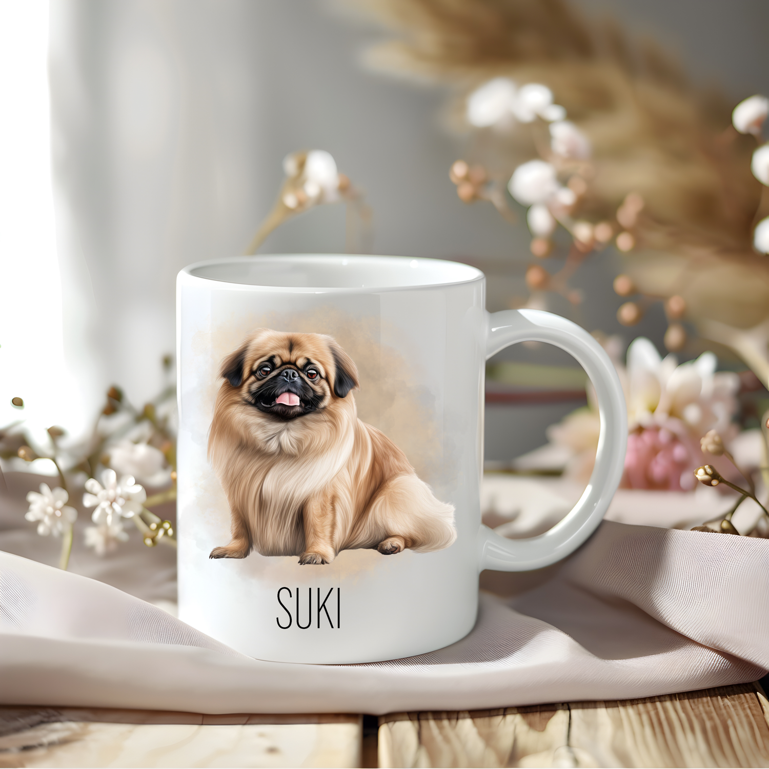 A Pekingese dog coffee mug personalised with a pet name.  The mug is sat on a wooden surface with a creamy coloured cloth and various flowers of pink and white adorn the background.