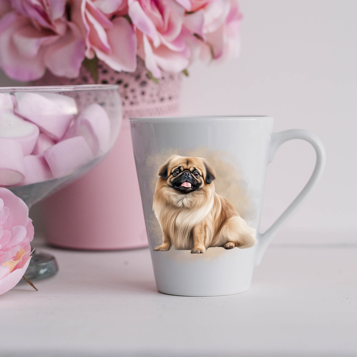 A Pekingese do is sitting on a white ceramic latte mug in a smoky background and looking forward.  The mug is placed on a white surface with pink flowers in a pink plant pot and a dish of pink marshmallows.