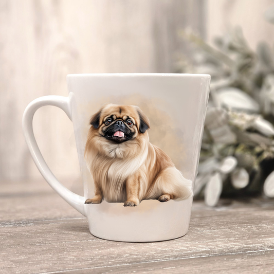 A Pekingese is shown sitting n a white ceramic latte mug in a smoky background and looking forward with mouth open and tongue out.  The mug is placed on a wooden surface with sage green foliage to the right-hand side.