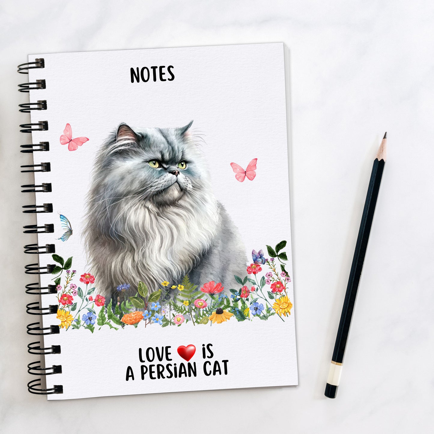 A white spiral bound notebook, with a grey Persian cat sat among pastel coloured wildflowers and butterflies flying around. A quote is seen at the bottom of the book and a pencil is laid to the side.