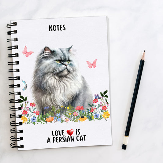 A white spiral bound notebook, with a grey Persian cat sat among pastel coloured wildflowers and butterflies flying around. A quote is seen at the bottom of the book and a pencil is laid to the side.