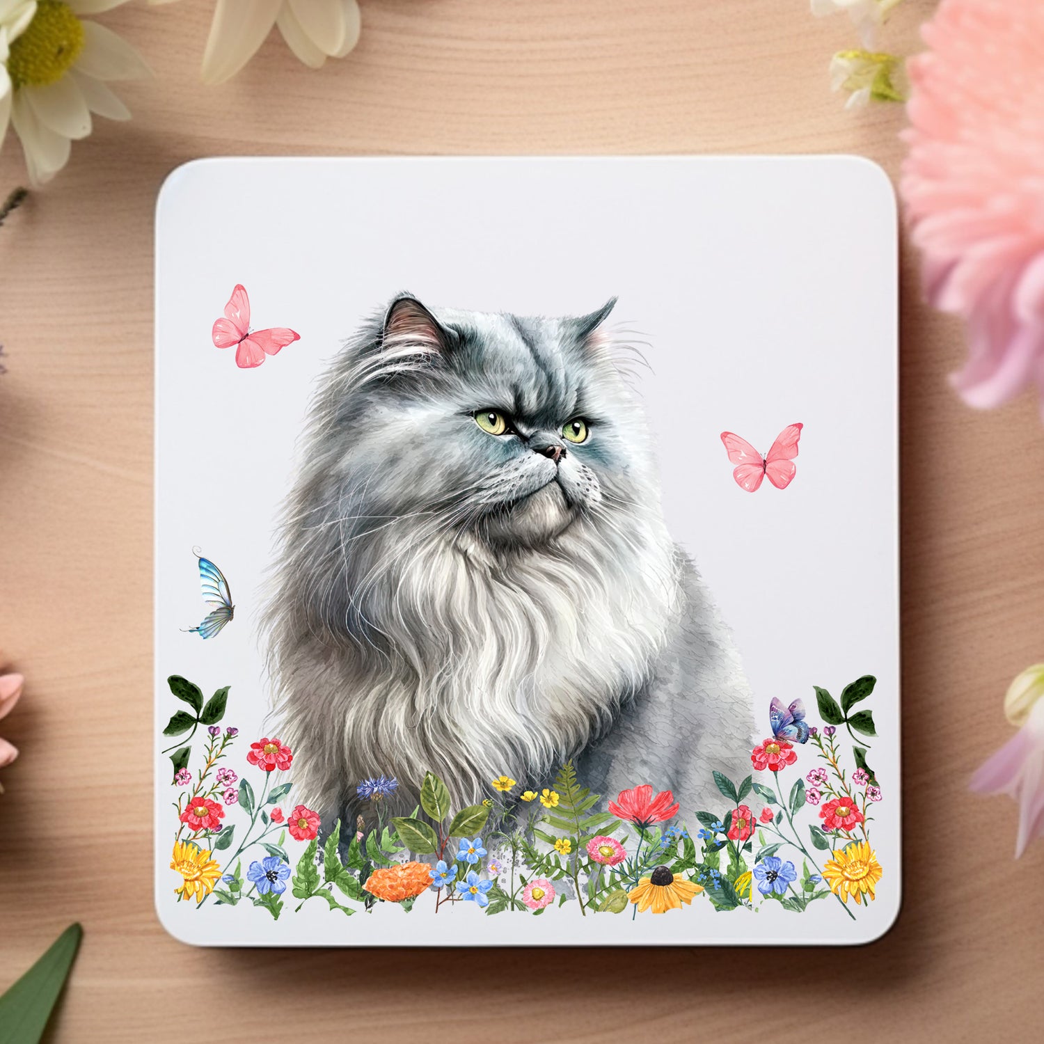 Grey Persian cat sat among flowers on a white coaster.  The coaster is on a wooden surface