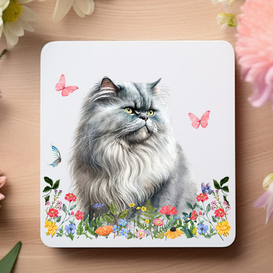 Grey Persian cat sat among flowers on a white coaster. The coaster is on a wooden surface
