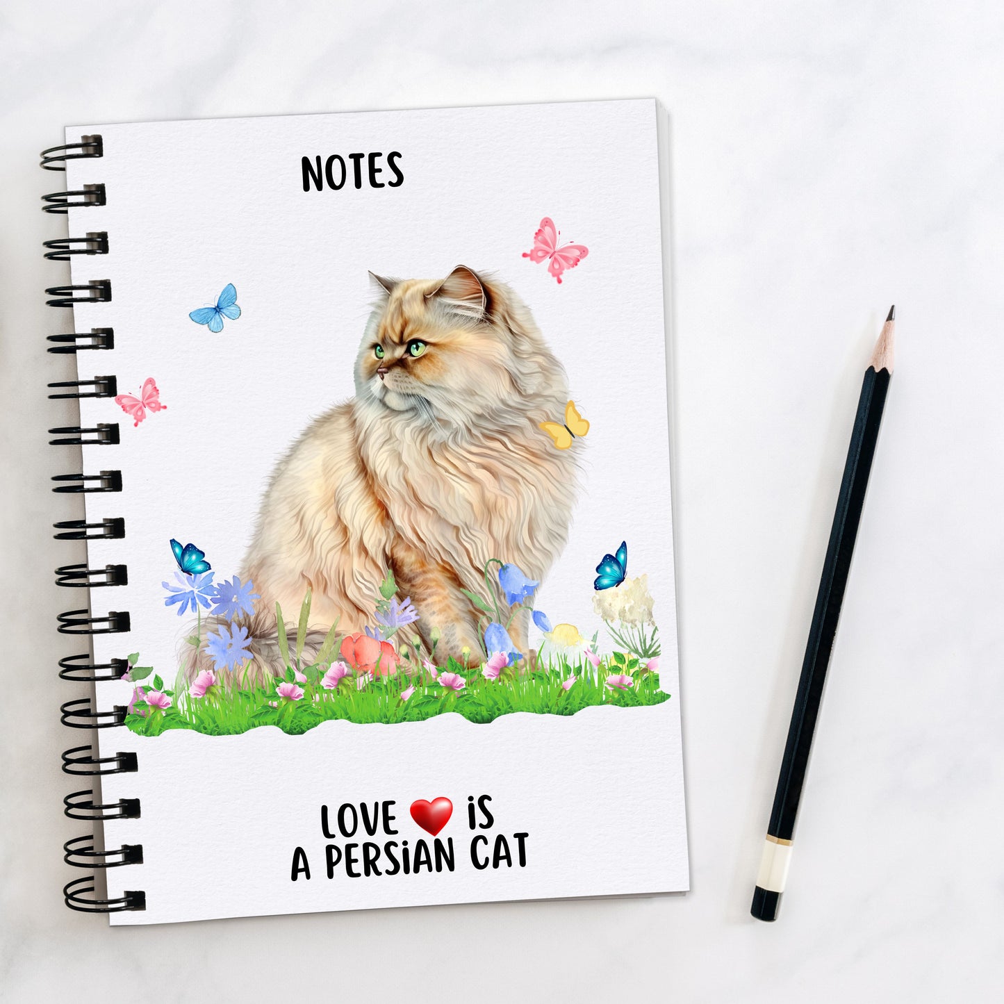 A white spiral bound notebook, with a ginger persian cat sat on grass among pastel colour flowers and butterflies flying around. A pencil is laid to the side.