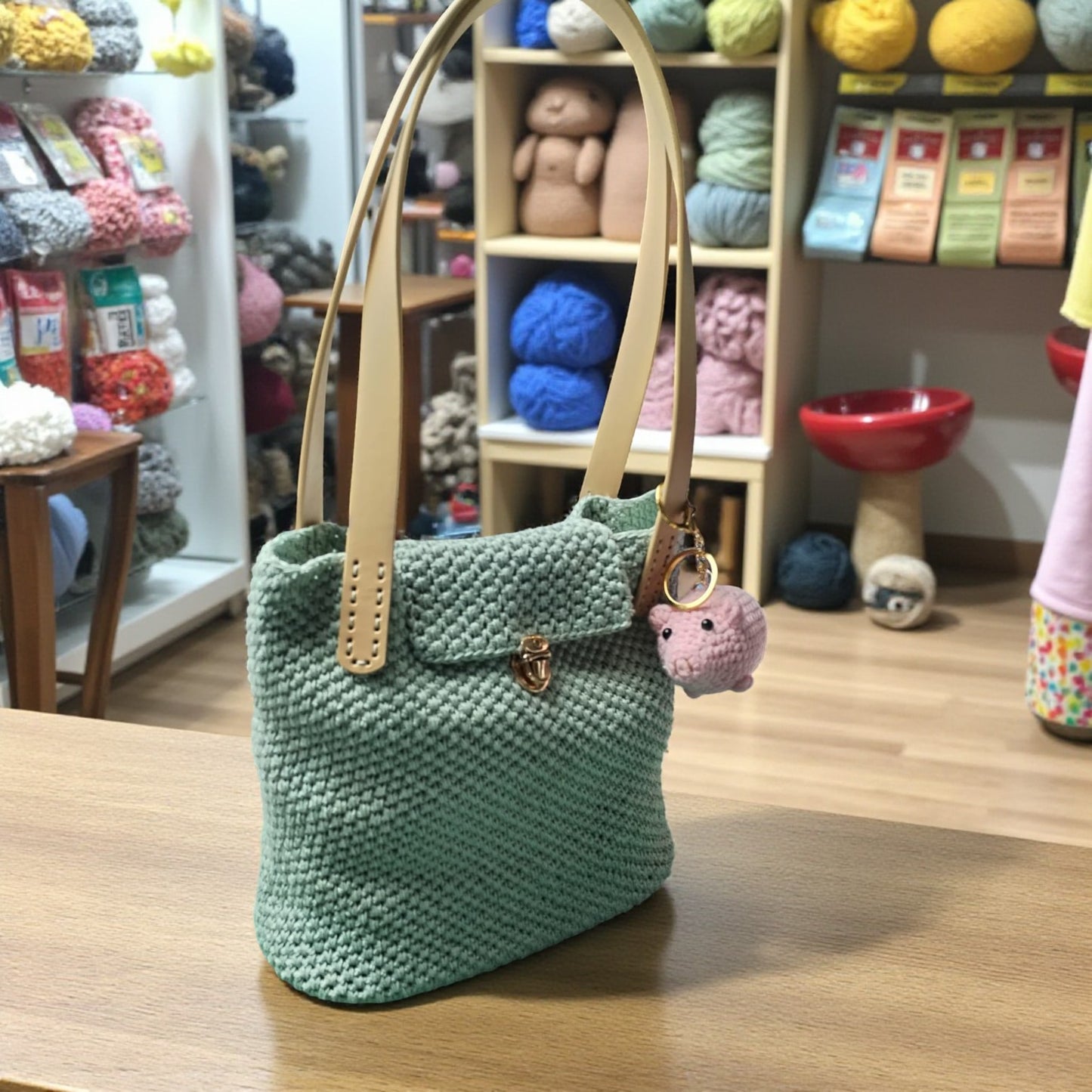 Green crochet    handbag with beige handle with a crochet pig key ring on a wooden surface, yarn and knitting supplies in the background.