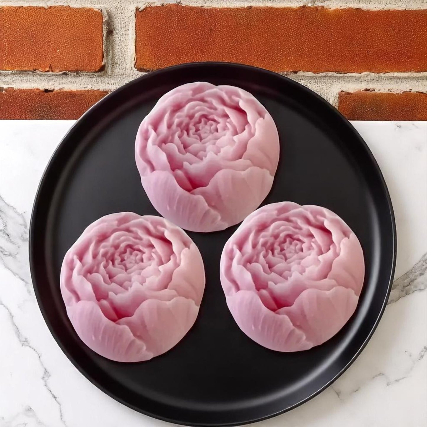 Three pink floral-shaped wax melts on a black plate with a brick wall background