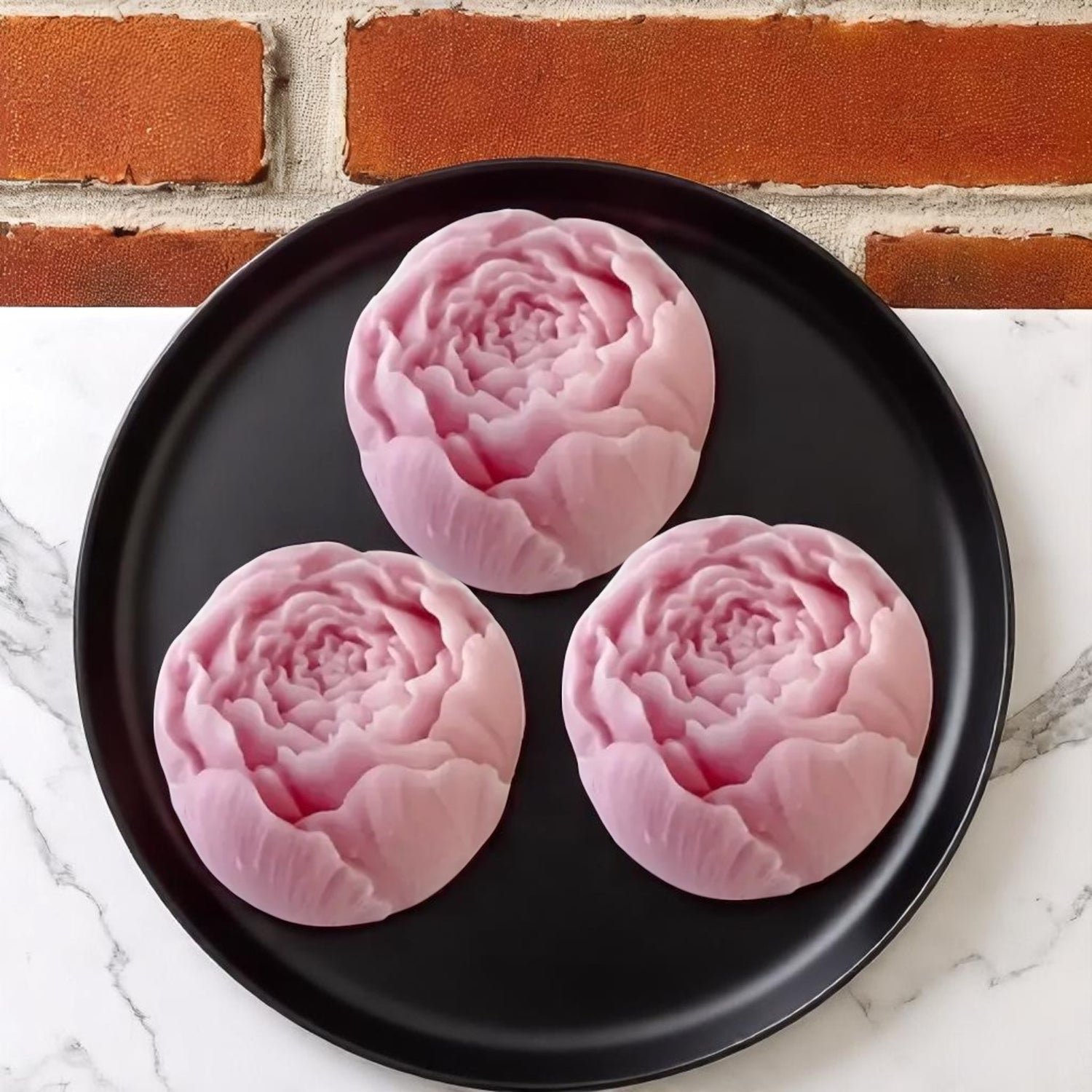 Three pink floral-shaped wax melts on a black plate with a brick wall background