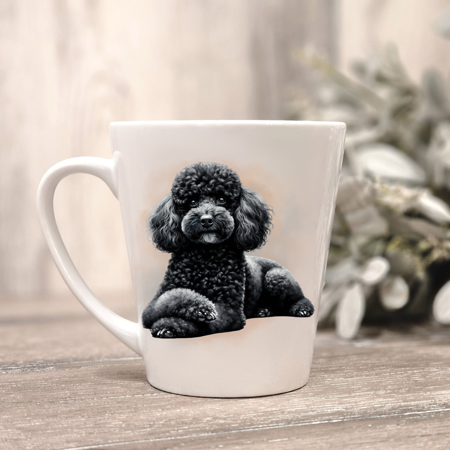 A black Miniature Poodle is shown on a white ceramic latte mug in a smoky background and looking forward.  The mug is placed on a wooden surface with sage green foliage to the right-hand side.