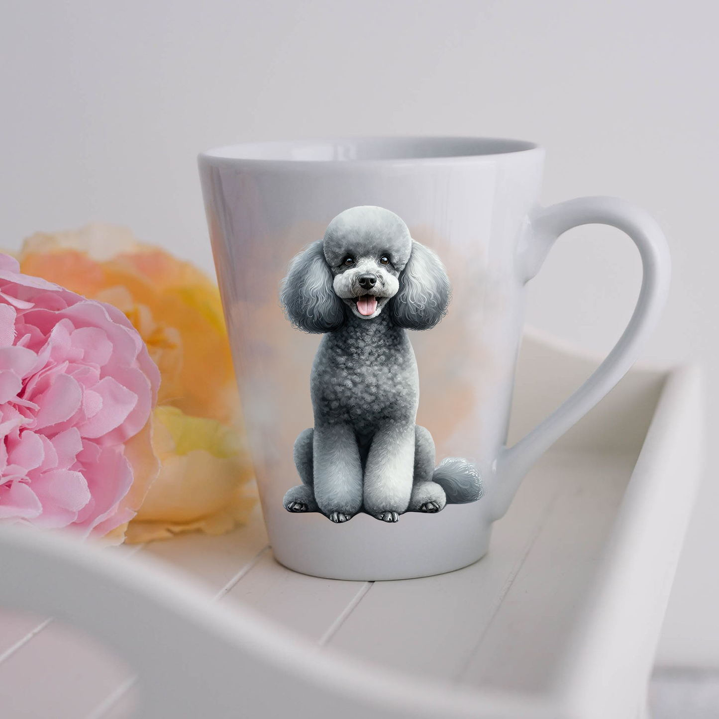 A grey Miniature Poodle is shown sitting on a white ceramic latte mug in a smoky background and looking forward with mouth open and tongue as if smiling.  The mug is placed on a white tray with pink and yellow flowers.