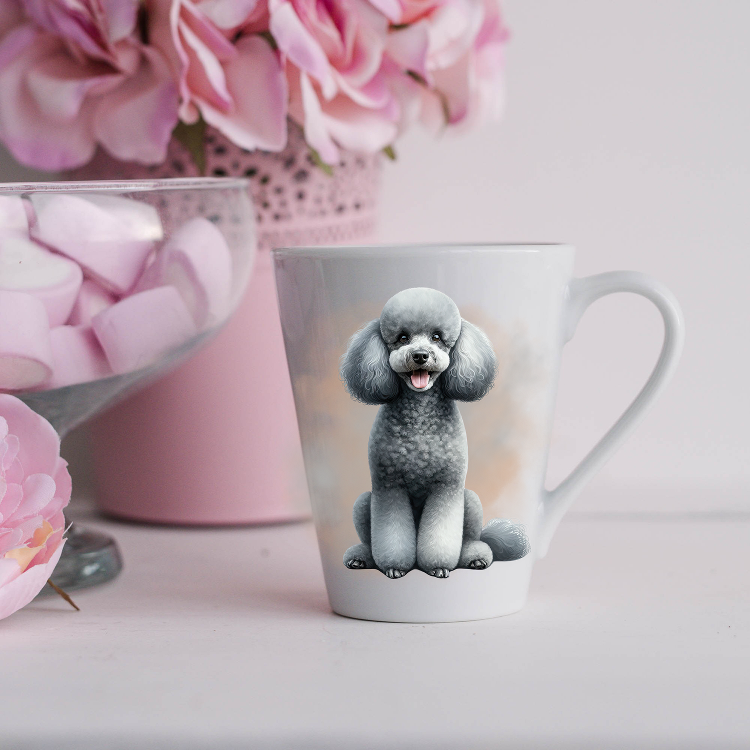 A Grey Miniature Poodle is shown on a white ceramic latte mug in a smokey background and looking forward.  The mug is placed on a white surface with pink flowers in a pink plant pot and a dish of pink marshmallows.
