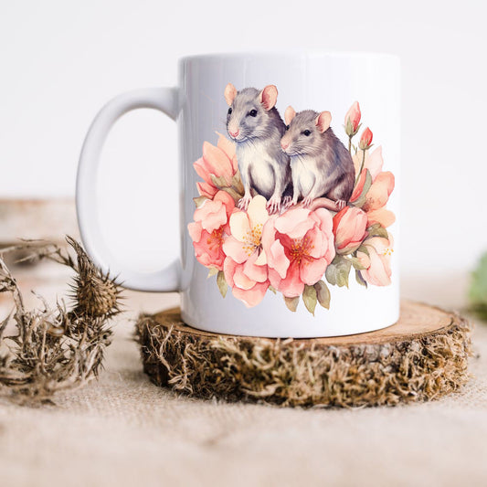 Two grey rats sat on pink and peach flowers shown on a white ceramic mug. The mug is placed on a rustic wooden disc.