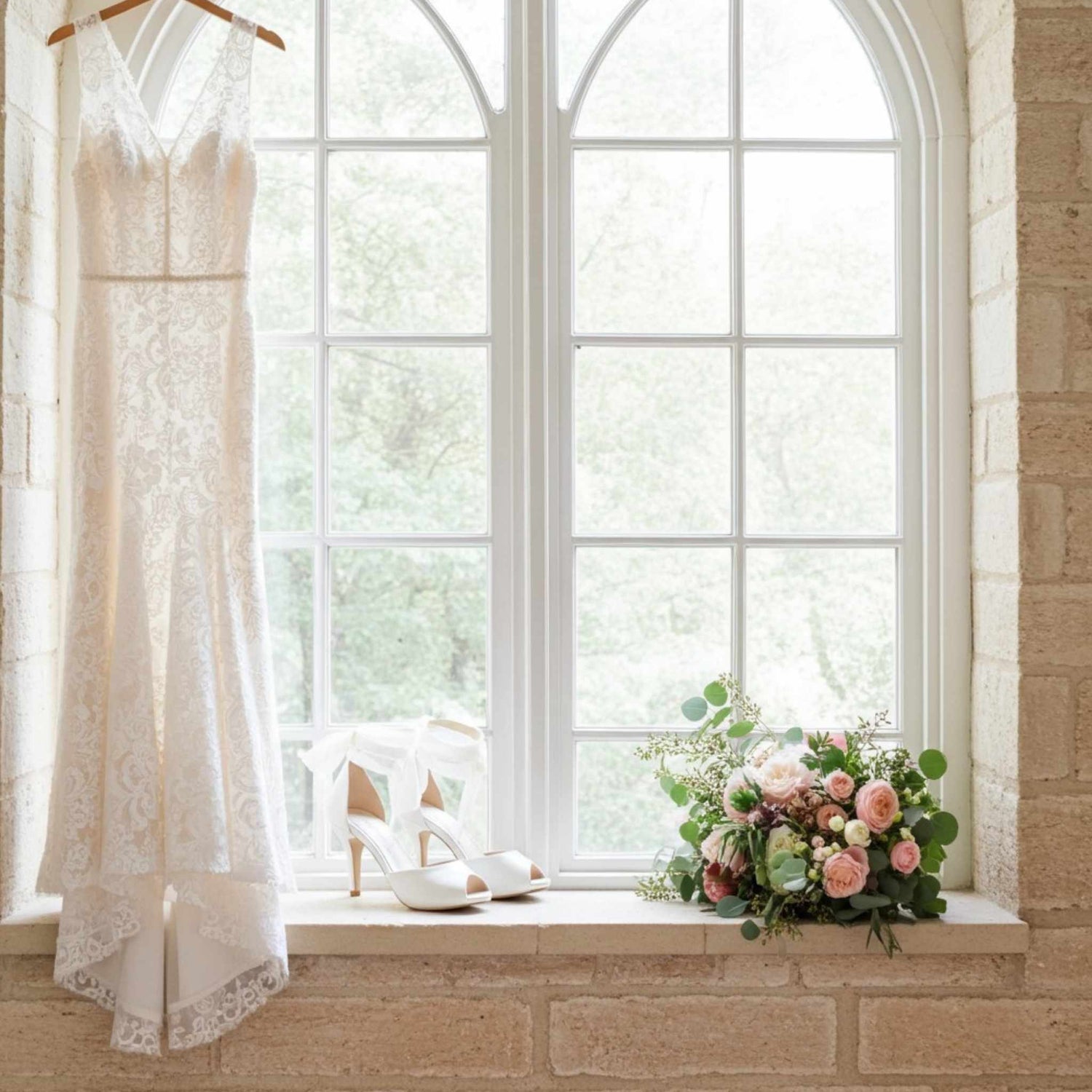 Lace wedding dress on a hanger, ivory peep toe high-heeled wedding shoes, and a bouquet of flowers on a windowsill.