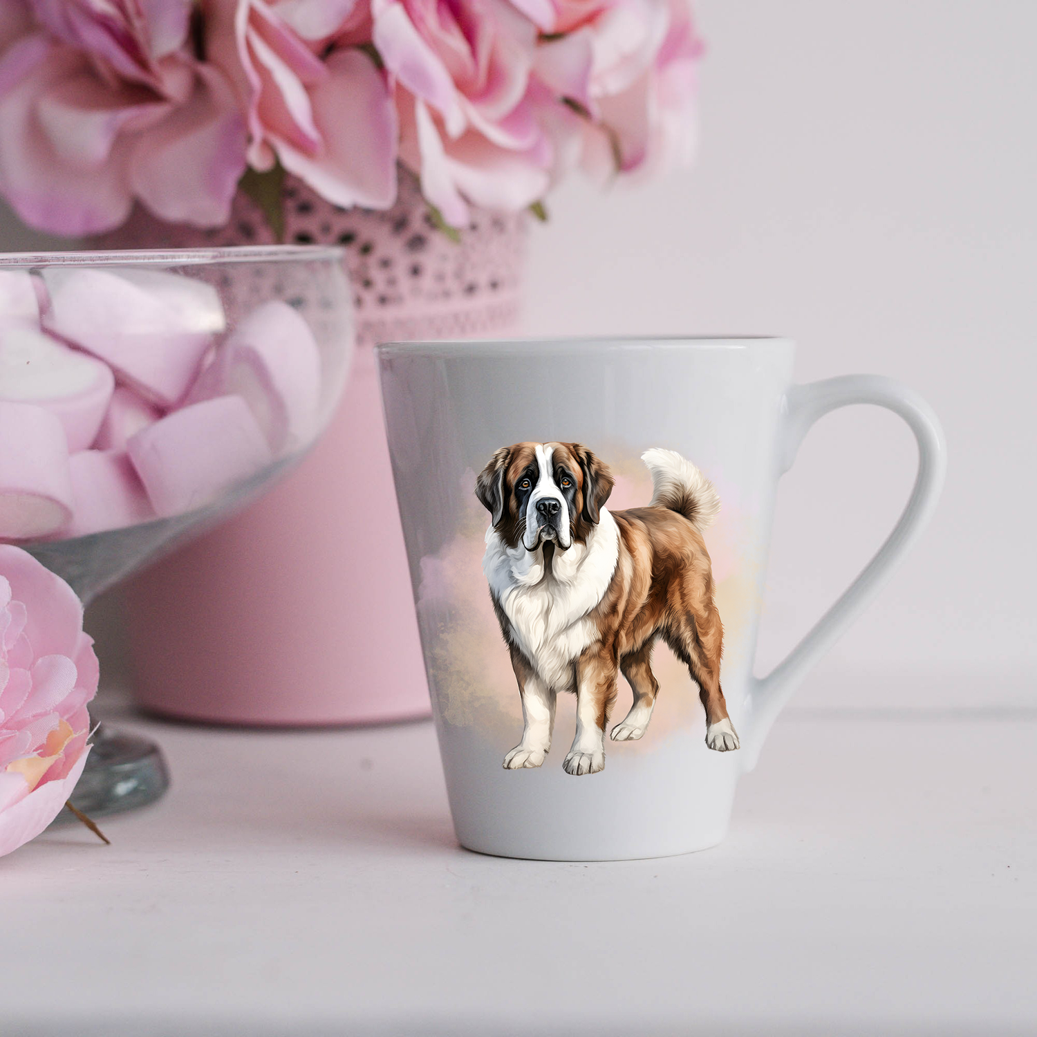 A Saint Bernard is shown standing on a white ceramic latte mug in a smoky background and looking forward.  The mug is placed on a white surface with pink flowers in a pink plant pot and a dish of pink marshmallows.