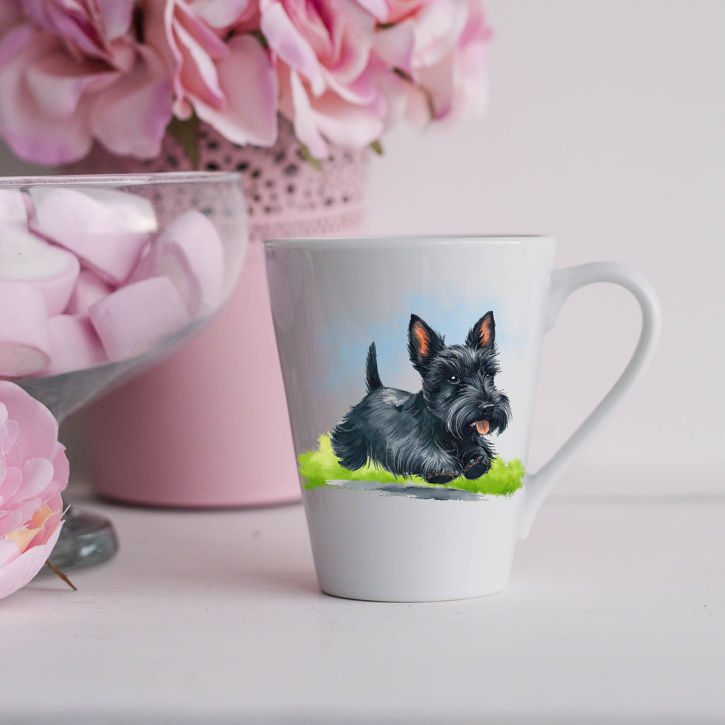 A black Scottish Terrier is running on a grassy surface with a blue sky on a white ceramic latte mug. The mug is placed on a white surface with pink flowers in a pink plant pot and a dish of pink marshmallows.