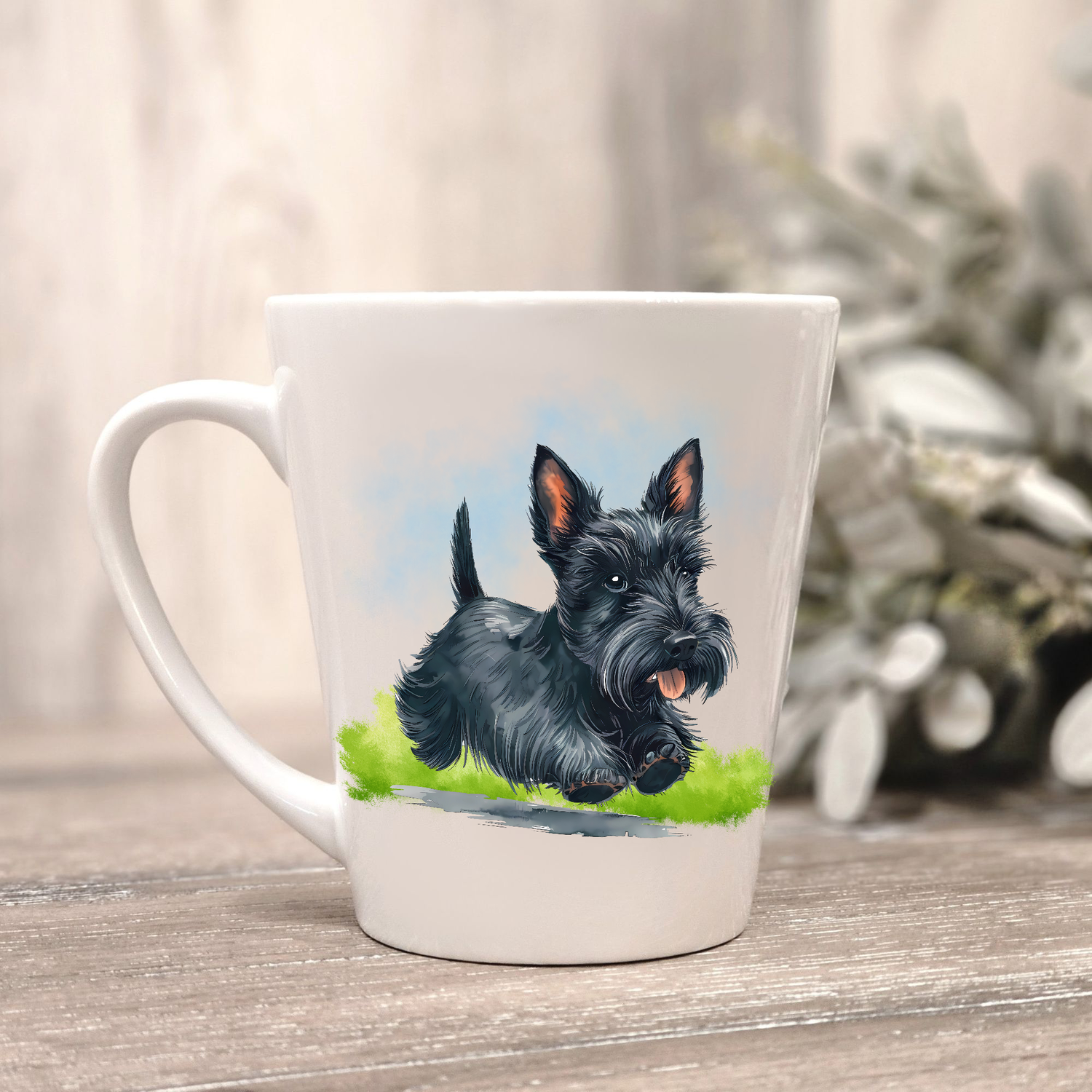 A black Scottish Terrier is running on a grassy surface under a blue sky on a white ceramic latte mug. The mug is placed on a wooden surface with sage green foliage to the right-hand side.
